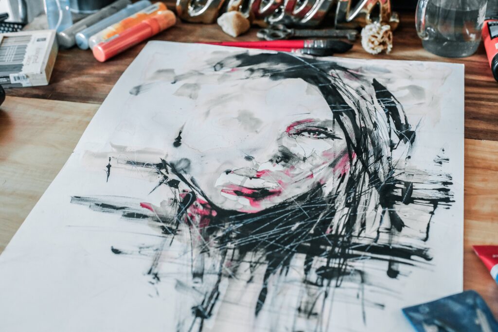 Photo of an artist's desk with pens & inks & paints visible around the edges of a paper flat on the desk with a faint outline of a woman's face where some of her hair and one eye and a little of her mouth are more complete but much of the painting still needs pieces done