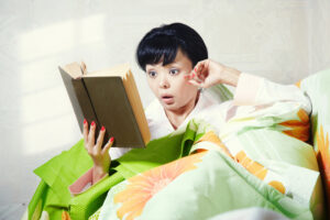 Photograph of a person reading a physical book in bed with eyes wide and mouth open
