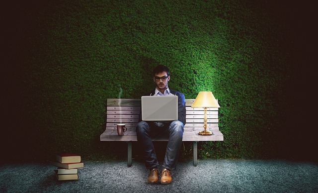 Man with a beard sits on a bench, typing on a laptop, with a cup of coffee on one side and a lamp on the other.